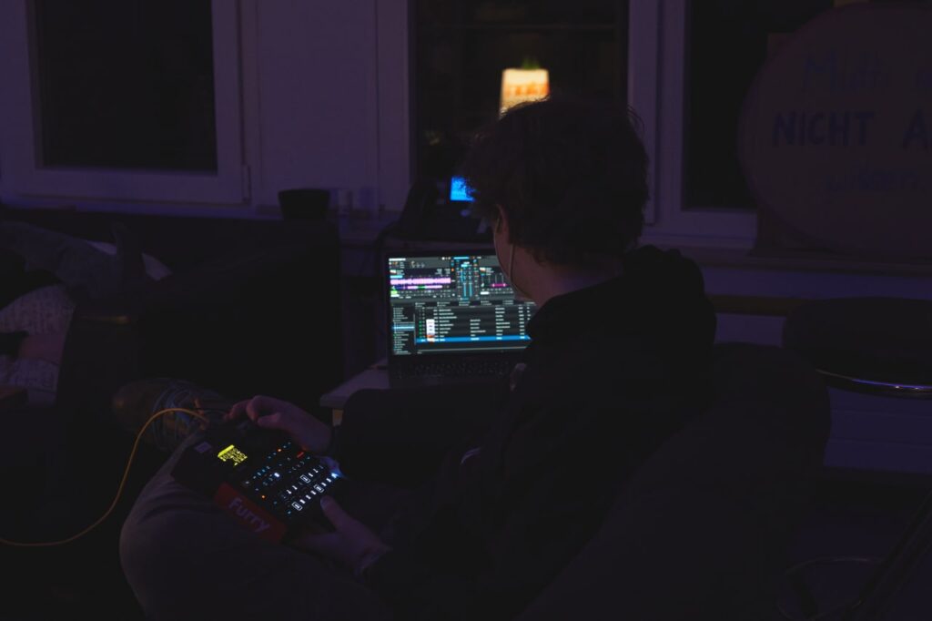Nik legt in der Lounge Musik auf. Auf einem Notebook ist das Interface einer DJ-Software zu sehen, auf seinem Schoß befindet sich ein Sampler bzw. Controller.