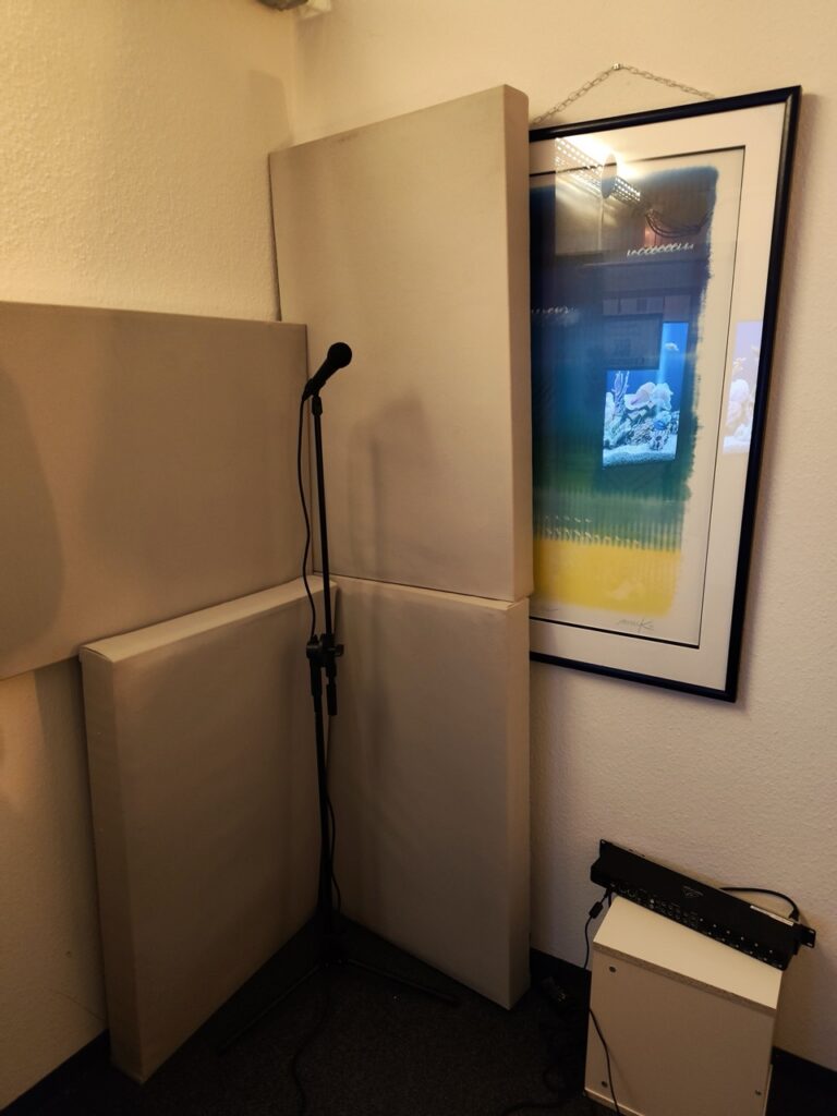 Mehrere, gegen eine Wand gestapelte Akustikpanele mit einem Mikrofon auf Stativ. Gemälde auf rechter Seite an der Wand.